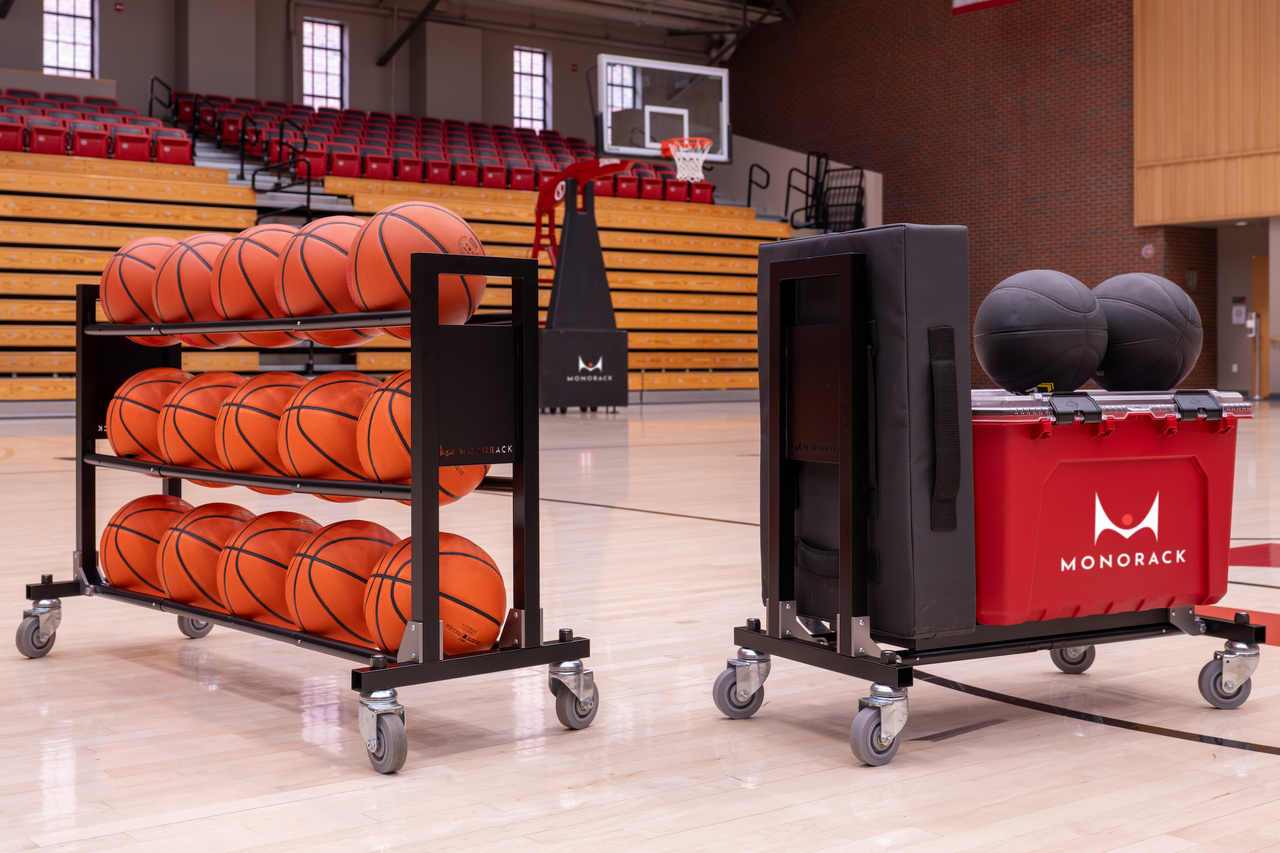 Two Monoracks situated on a basketball court. One ball rack  is collapsed and has equipment stacked on it, the other is loaded with 15 basketballs.