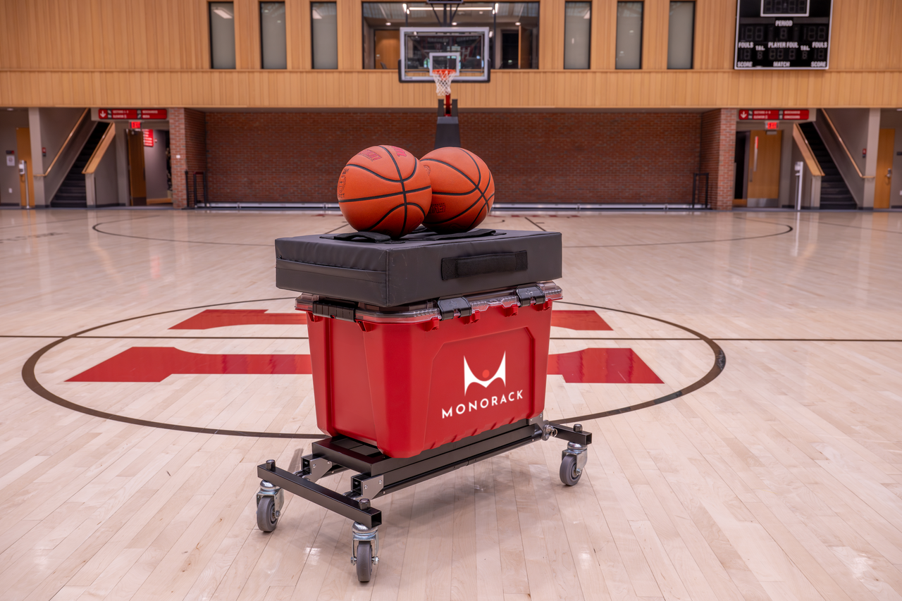 A Monorack with a bin, a training pad, and two basketballs stacked on top of it in an empty gym.